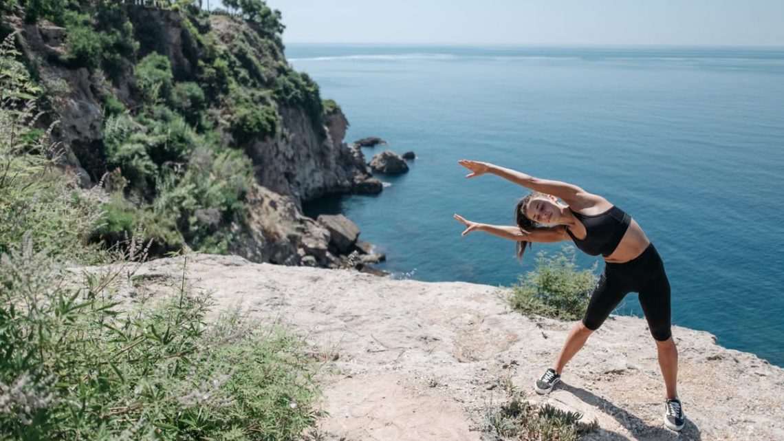 A woman in activewear stretches on a cliff by the ocean, embracing coastal fitness.