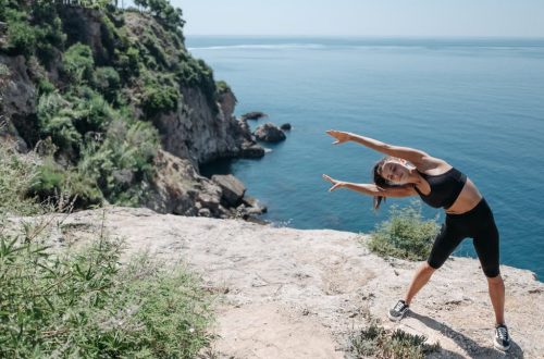 A woman in activewear stretches on a cliff by the ocean, embracing coastal fitness.