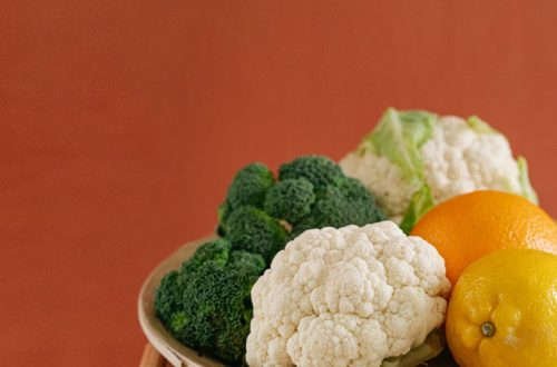 Close-up of fresh broccoli, cauliflower, lemon, and orange on a wooden plate with an earthy background.